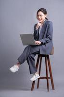 Young Asian businesswoman sitting on chair and using laptop on gray background photo