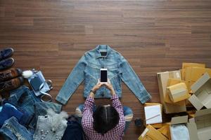 vista superior de las mujeres toman una foto de una chaqueta de jeans con un teléfono inteligente desde casa en un piso de madera con un paquete postal, vendiendo el concepto de ideas en línea