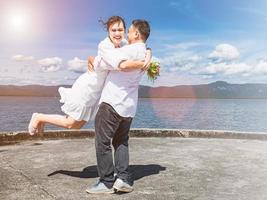 An Asian woman wearing a white dress holding a bouquet of flowers happily hugs an Asian man at the sea port. To show love after being proposed in the sunshine, clear sky, strong wind