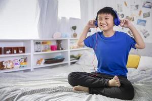 un chico asiático con auriculares azules está usando un teléfono inteligente y sonriendo feliz mientras escucha música en la cama en casa foto