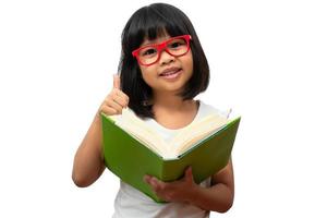 Happy Asian little preschool girl wearing red glasses holding a green book and thumbs up on white isolated background. Concept of school kid and education in elementary and preschool, home school