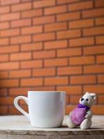 oso de peluche sosteniendo un corazón morado. oso de peluche junto al café con leche colocado sobre un escritorio de madera. el telón de fondo es un bloque de ladrillo de color marrón. foto