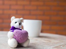 oso de peluche sosteniendo un corazón morado. vivir en una taza blanca de café. colocado sobre un escritorio de madera. el telón de fondo es un bloque de ladrillo de color marrón. foto