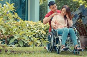 Asian woman in a wheelchair and Unhappy and painful. A Man standing behind the wheelchair and is encouraging his wife, whose feet hurt her leg due to an accident. Concept of caring and support photo