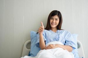 feliz paciente asiática sonriendo, se acuesta en la cama y levanta el pulgar para mostrar confianza en el tratamiento. concepto de creer en el tratamiento y la cobertura de seguro foto