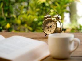 text book with vintage alarm clock and White coffee cup on the wood table. The background is green from tree and light bokeh photo