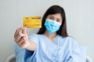 Happy Asian woman wearing a medical mask and holding mock up credit card insurance card and ok sign in a hospital bed. Insurance policy by bank, payment medical treatment concept photo