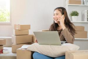 Happy Asian young entrepreneur, talking on the smartphone with customer for tack order, She smile for sales success at home office, Concept of merchant business online and eCommerce photo