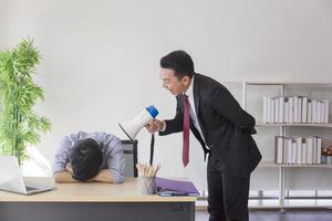 un supervisor asiático usa un megáfono para gritar a sus subordinados dormidos en una mesa de oficina.