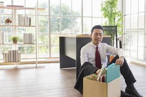 An Asian male company worker sits on the floor beside a desk with a cardboard box containing his personal belongings. photo