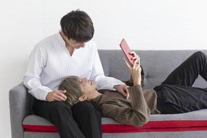 A male couple with an Asian man sitting on a sofa with their love for one another and one playing tablet. photo