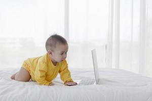 un bebé asiático está jugando en una computadora portátil en una cama blanca y limpia. foto