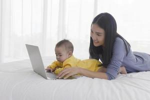 una madre asiática está trabajando en una computadora portátil en la cama y con su bebé junto al concepto de trabajo en casa. foto