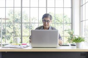 A middle-aged Asian man holding a laptop in his hand sits on a desk beside a glass panel, taking a video call photo