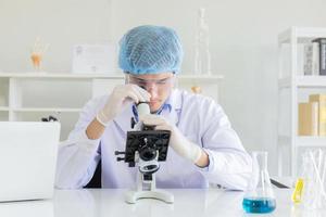 Young Scientist using Microscope in Laboratory. Male Researcher wearing white Coat sitting at Desk and looking at Samples by using Microscope in Lab. Scientist at Work in Laboratory photo