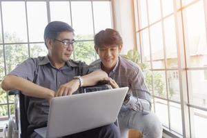 A middle-aged Asian father sits in a wheelchair and has a son next to him chatting on video calls on a laptop. photo