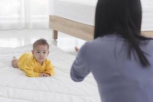 An Asian infant who is practicing crawling With her mother sitting on the other side of her soft white cloth in the bedroom photo