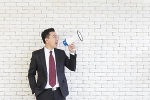 An Asian man in a suit holds a megaphone on a white brick wall. photo