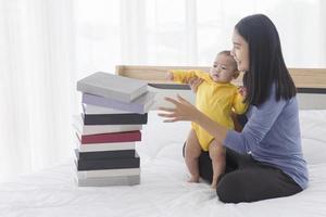 An Asian mother helped her baby to stand. And there was a stack of books placed next to her on the bed. photo