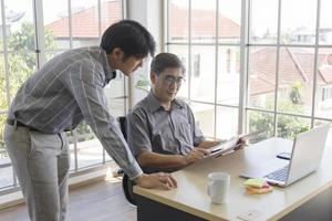 A middle-aged Asian man teaching his son a job standing next to a desk in an office. photo