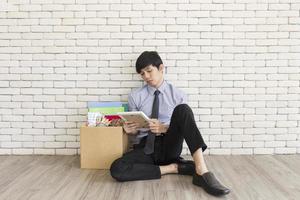 An Asian man sat sadly on a wooden floor in an office after being fired, storing personal belongings in cardboard boxes. photo