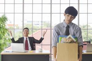 un hombre asiático de pie tristemente sosteniendo una caja de cartón que contiene sus pertenencias personales y su gerente sonriendo detrás de él en un escritorio en una oficina. foto