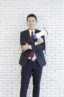 An Asian man in a suit holds a megaphone on a white brick wall. photo