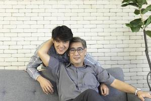 A middle-aged father and an Asian son hugged each other on the sofa with a happy smile. photo