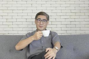 An Asian middle-aged man holding a coffee cup is sipping on the sofa. photo