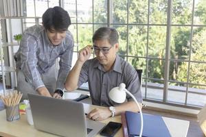 A middle-aged Asian man teaching his son a job standing next to a desk in an office. photo