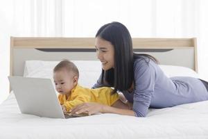 An Asian mom is working on a laptop in bed and with her baby next to the concept of work at home. photo