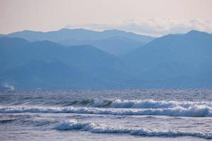 Stormy sea waves hit a sandy beach. Snow capped mountains appear in the distance. photo