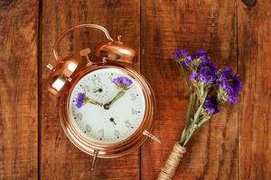 gold alarm clock with blue flowers instead of clock's hands and small bouquet of flowers on wooding surface. Spring time photo