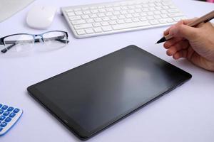 la tableta de pantalla blanca en blanco y los dispositivos asociados rodean una imagen de vista superior de una estación de trabajo blanca.