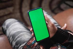 mano de un joven usando un teléfono inteligente con pantalla verde sentado en un sofá foto