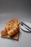 Pile of sausage breads and calzone pizza on a wooden cutting board. Side view of home made sausage breads and calzone pizza in a light grey background with knife and fork on a white plate photo