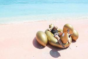 Asian woman in summer hat and sunglasses laying on inflatable swan in pink sandy beach photo