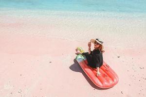 Blondde woman in summer hat and sunglasses smiling at camera while sitting on inflatable mattress on pink sandy beach