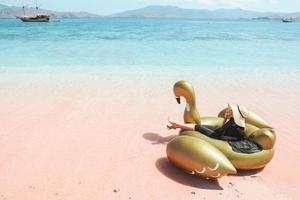 Female tourist in summer hat relaxing on golden inflatable swan on pink sandy beach