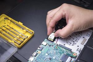enfoque selectivo en la mano del técnico que sostiene el chip de la computadora para instalar en la placa base del circuito electrónico de la computadora portátil con una caja de destornilladores amarilla en la mesa negra foto