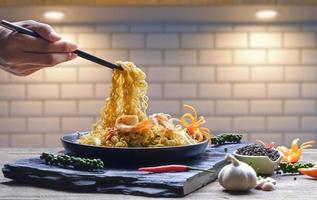 Man hand using chopsticks to picking up stir fry instant noodles spicy seafood on black ceramic plate in the kitchen photo