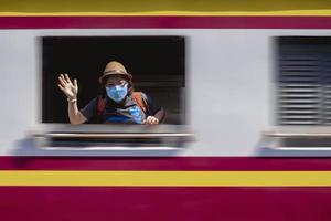 movimiento borroso de una viajera con máscara protectora mirando la cámara y agitando la mano desde la ventana del tren mientras el tren sale de la estación foto