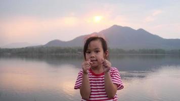 una niña linda sonríe y mira la cámara durante la puesta de sol junto al lago. viajar en vacaciones de verano con la familia. video