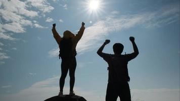 Silhouette of a successful Asian couple raising their arms on top of a mountain enjoying the valley view before sunset. video