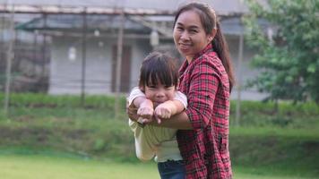 Happy Asian mother holds her daughter in her hands, little daughter raises her hand, imagining herself as a plane in the summer garden. video
