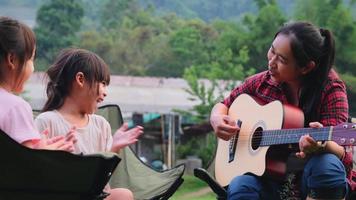 moeder en kind spelen gitaar en zingen samen op campingstoelen in de buurt van tent op kamp in zomerbos. het gezin brengt samen tijd door op vakantie. video