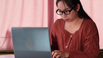 mooie aziatische vrouw in vrijetijdskleding gebruikt een laptop terwijl ze binnenshuis werkt. jonge zakenvrouw met een bril die op het werk zit en op een laptop typt. video