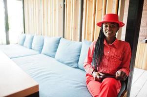 Pretty braids business african american lady bright bossy person friendly wear office red shirt, hat and trousers, hold tablet on hands. photo