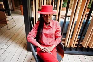 Pretty braids business african american lady bright bossy person friendly wear office red shirt, hat and trousers, sit on chair.