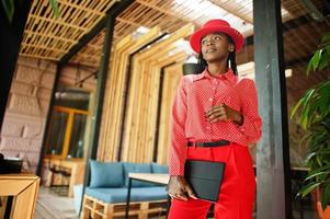 Pretty braids business african american lady bright bossy person friendly wear office red shirt, hat and trousers, hold tablet on hands. photo